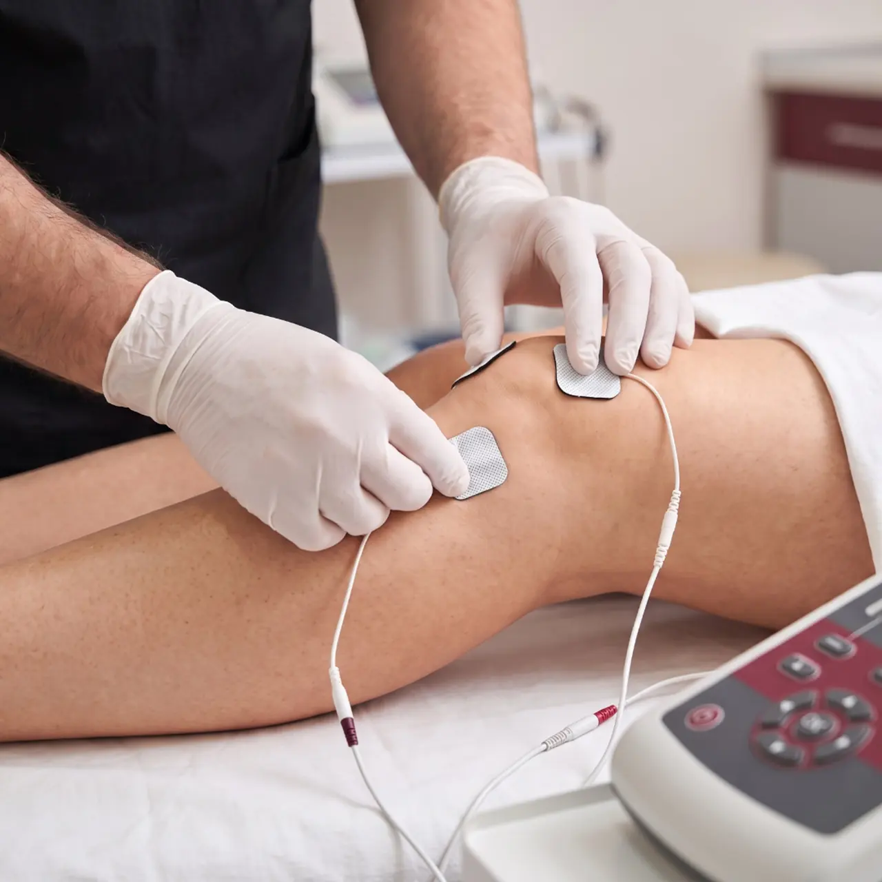 Nerve stimulation setup Gloved hands placing electrode pads on a patient's knee beside a stimulation unit